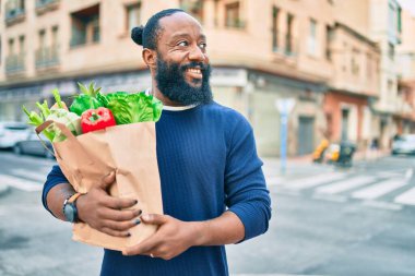 Sakallı Afrikalı Amerikalı bir adam elinde süpermarketten alınmış kağıt poşetler tutuyor.