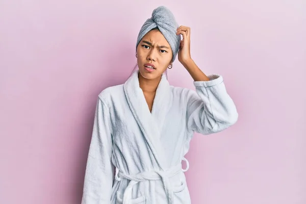 Young chinese woman wearing shower towel cap and bathrobe confuse and wonder about question. uncertain with doubt, thinking with hand on head. pensive concept.