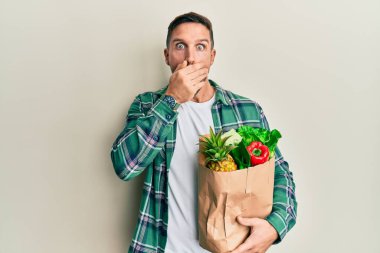 Elinde alışveriş poşeti olan sakallı yakışıklı bir adam ağzını yanlışlıkla elleriyle kapatmış. gizli kavram. 