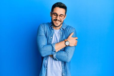 Gündelik kıyafetler ve gözlük takan genç İspanyol adam kendine sarılıyor mutlu ve pozitif, kendinden emin gülümsüyor. Sevgi ve özgüven. 