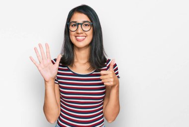 Güzel Asyalı genç bir kadın günlük kıyafetler ve gözlük takıyor. Altıncı parmaklarını gösterip işaret ediyor. Kendinden emin ve mutlu bir şekilde gülümsüyor.. 
