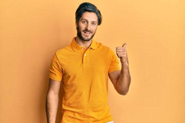 Günlük sarı tişört giyen, gülümseyen, gülümseyen ve başparmağıyla yan tarafı gösteren İspanyol genç adam.. 