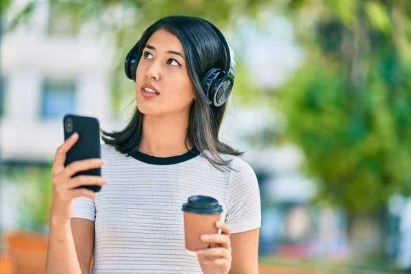 若いヒスパニック系の女性がコーヒーを飲み 市内でスマートフォンやヘッドフォンを使用して — ストック写真