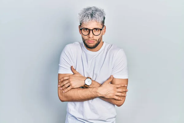 Young hispanic man with modern dyed hair wearing white t shirt and glasses shaking and freezing for winter cold with sad and shock expression on face