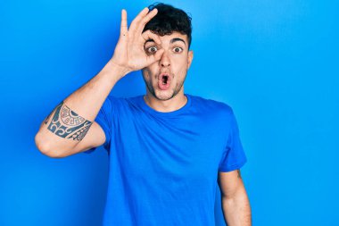 Latin asıllı genç bir adam günlük mavi tişört giyiyor. Şaşırmış yüz ifadesiyle şaşırmış, parmaklarının arasından bakıyor. İnançsız ifade. 