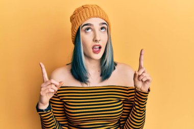 Yün şapka takan genç modern kız hayret ve şaşkınlık içinde yukarı bakıyor ve parmaklarıyla işaret ediyor ve kollarını kaldırıyor.. 