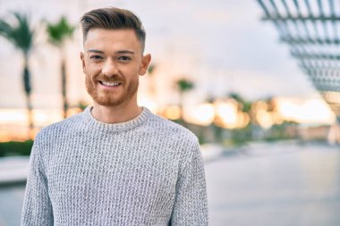 Genç beyaz adam gülümseyerek şehirde mutlu mesut duruyor..