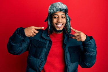 Sakallı, kışlık şapkalı ve ceketli neşeli gülümseyen ve parmaklarıyla ve ağzıyla işaret eden Afrikalı genç bir Amerikalı. diş sağlığı kavramı. 