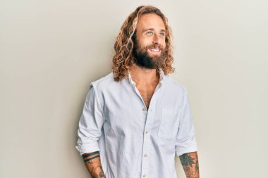 Sakallı, uzun saçlı, sıradan beyaz tişörtlü, rahat bir görünüm takınan, kendine güvenen ve gülümseyen yakışıklı bir adam.. 