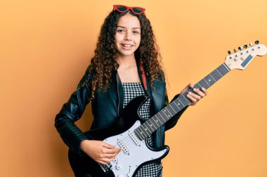 Elektro gitar çalan genç İspanyol kız mutlu ve havalı bir gülümsemeyle gülümsüyor. diş gösterme. 