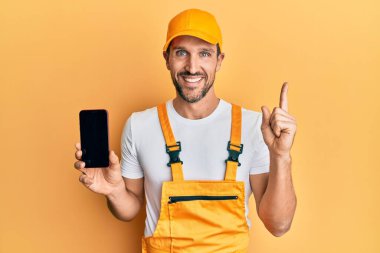 Becerikli üniformalı genç adam akıllı telefon ekranı gösteriyor. Gülümsüyor, mutlu bir şekilde elini ve parmağını yana doğru gösteriyor. 