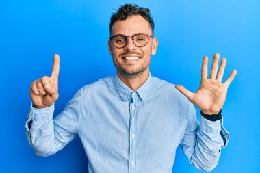 Gündelik kıyafetler giyen ve gözlük takan İspanyol genç adam altı numarayı gösterip işaret ederken kendinden emin ve mutlu gülümsüyor.. 