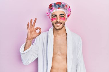 Bornoz ve duş başlığı takan genç İspanyol adam göz bantları kullanıyor. Parmaklarıyla imza atıyor, gülümseyerek mükemmel bir sembol oluşturuyor. 