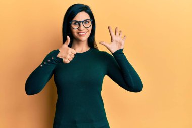 Genç beyaz kadın, günlük kıyafetler ve gözlük takıyor. Altıncı parmaklarını gösterip işaret ediyor. Kendinden emin ve mutlu bir şekilde gülümsüyor.. 