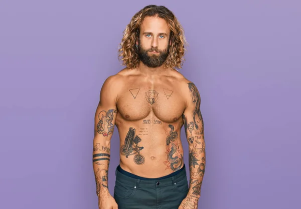 Handsome man with beard and long hair standing shirtless showing tattoos looking sleepy and tired, exhausted for fatigue and hangover, lazy eyes in the morning.