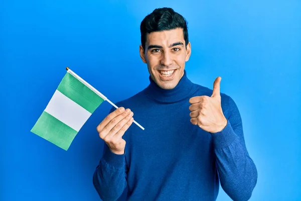Hombre Hispano Guapo Sosteniendo Bandera Nigeria Sonriendo Feliz Positivo Pulgar —  Fotos de Stock
