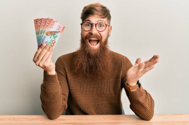 Genç İrlandalı kızıl saçlı adam elinde 100 yeni Zelanda Doları banknotuyla masaya oturmuş başarıyı mutlu bir gülümsemeyle ve kazanan yüz ifadeyle kutluyor. 