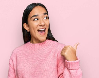 Genç Asyalı kadın, günlük kışlık süveter giyiyor, gülümsüyor, gülümsüyor ve başparmağıyla yan tarafa bakıyor.. 