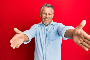 Orta yaşlı, gri saçlı, sıradan kıyafetler giyen, kameraya bakıp sarılmak için kollarını açmış gülümseyen bir adam. Mutluluğu kucaklayan neşeli bir ifade.. 