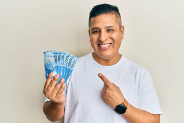 Young latin man holding 10000 chilean pesos smiling happy pointing with hand and finger  clipart