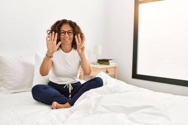 Orta yaşlı İspanyol bir kadın evinde yatağında oturmuş 8 numaralı parmaklarıyla işaret ederken kendinden emin ve mutlu bir şekilde gülümsüyor.. 