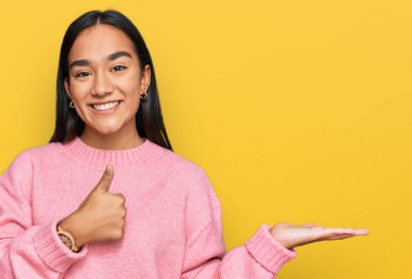 Genç Asyalı kadın, günlük kışlık süveter giyiyor avuç içi gösteriyor ve baş parmaklarıyla iyi bir jest yapıyor, mutlu ve neşeli gülümsüyor. 