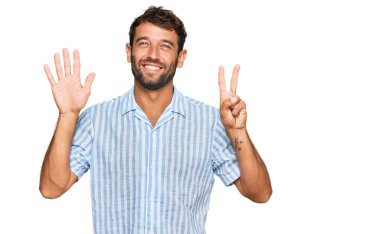 Sakallı yakışıklı genç adam, günlük kıyafetlerini giyiyor ve yedi numaralı parmaklarıyla işaret ediyor. Kendinden emin ve mutlu bir şekilde gülümsüyor.. 