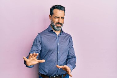 Orta yaşlı, sakallı, gri saçlı, iş kıyafetleri giyen, tiksindirici ve tiksindirici bir yüz ifadesi takınan bir adam. Eller havada. 