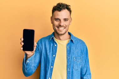 Akıllı telefonu tutan genç İspanyol adam boş ekranı gösteriyor. Pozitif ve mutlu görünüyor. Kendine güvenen bir gülümsemeyle dişlerini gösteriyor. 