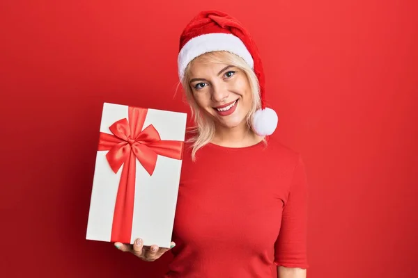 Menina Loira Jovem Usando Chapéu Natal Segurando Presente Olhando Positivo — Fotografia de Stock