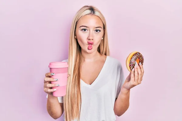 年轻的金发姑娘吃着甜甜圈 喝着咖啡 用嘴和眼神做鬼脸 疯狂而滑稽 — 图库照片