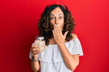 Orta yaşlı İspanyol kadın elinde kavanozla balkabağı çekirdeği ile ağzını kapatıyor, şok geçiriyor ve hata yapmaktan korkuyor. şaşırmış ifade 