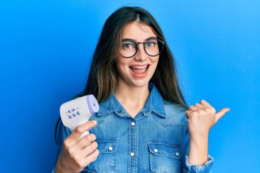 Dijital termometreyi tutan beyaz bir kadın baş parmağını yukarı kaldırıyor ve ağzı açık gülümsüyor. 