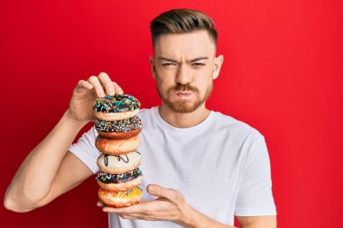 Genç kızıl saçlı adam elinde bir yığın lezzetli renkli donut tutuyor komik suratlı yanakları şişiriyor. Ağzı havayla şişer, havayı yakalar.. 