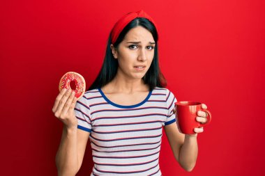 Genç İspanyol kadın donut yiyor ve kahve içiyor habersiz ve kafası karışık bir ifade. şüphe kavramı. 