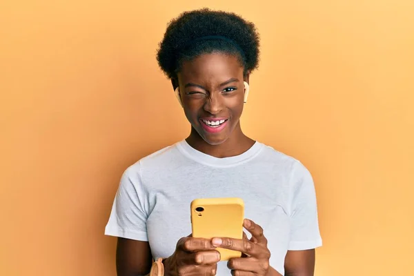 年轻的非洲裔美国女孩用智能手机和耳机眨眼 用性感的表情 开朗而快乐地看着摄像机 — 图库照片
