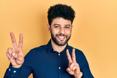 Sakallı, sıradan tişörtlü, gülümseyen, parmaklarını zafer işareti yaparken gösteren genç bir Arap. İki numara.. 