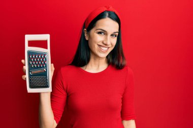 Genç İspanyol kadın elinde rendeleyici, pozitif ve mutlu görünüyor. Kendine güvenen bir gülümsemeyle ayakta duruyor ve dişlerini gösteriyor. 