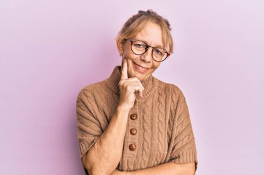 Pembe arka planda gözlük takan orta yaşlı sarışın kadın kameraya kollarını kavuşturup çenesine dokunarak gülümsüyor. Olumlu düşünmek. 