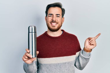 Sakallı yakışıklı beyaz adam termo tutuyor, gülümsüyor mutlu bir şekilde elini ve parmağını yana uzatıyordu. 