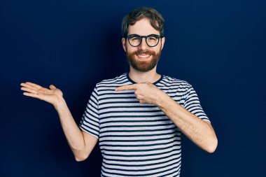 Beyaz sakallı, çizgili tişörtlü ve gözlüklü bir adam kameraya hayret ediyor ve gülümsüyor.. 