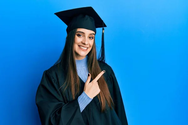 美しいブルネット若い女性が卒業キャップと式のローブを身に着けています陽気に笑顔で顔を指します手と指で側まで幸せと自然な表情で顔 — ストック写真