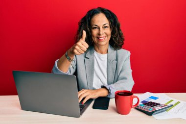 Ofiste çalışan orta yaşlı güzel bir kadın bir fincan kahve içerek mutlu bir el hareketi yapıyor. Kameraya bakıp başarıyı gösteren ifade. 