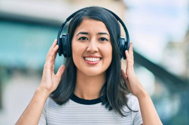 Genç İspanyol kadın gülümseyerek şehirde kulaklık takarak müzik dinliyor..