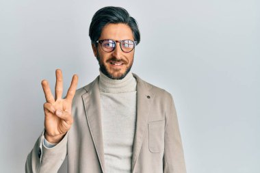 İş ceketi ve gözlük takan İspanyol genç adam üç numaralı parmaklarını gösterip işaret ederken kendinden emin ve mutlu gülümsüyor.. 