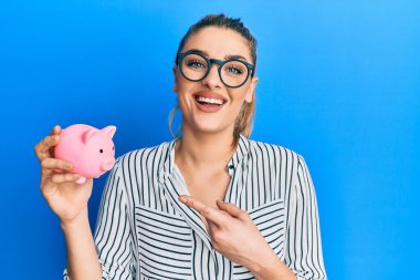 İş kıyafetleri giymiş genç beyaz bir kadın kumbarayı tutarken mutlu bir şekilde elini ve parmağını işaret ediyor. 