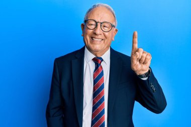 Beyaz üst düzey bir adam iş elbisesi ve kravat takıyor. Kendini beğenmiş ve mutlu bir şekilde gülümserken bir numaralı parmağını gösterip işaret ediyor.. 