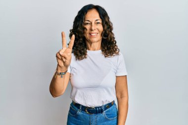 Orta yaşlı İspanyol kadın, günlük beyaz tişört giyer ve parmaklarıyla iki numarayı gösterirken kendinden emin ve mutlu bir şekilde gülümser.. 