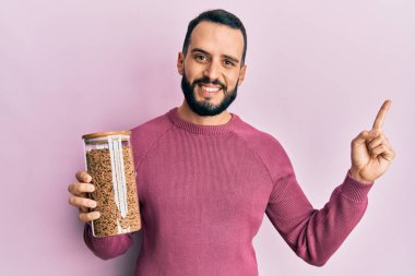 Sakallı genç adam elinde sağlıklı tahıl gevreği kavanozuyla gülümsüyor. 