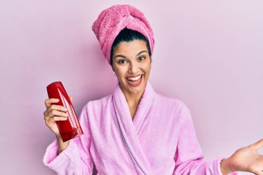 Duş bornozu giyen genç İspanyol kadın elinde şampuanla zaferi kutluyor mutlu bir gülümsemeyle ve elini kaldırarak kazanan bir ifadeyle. 